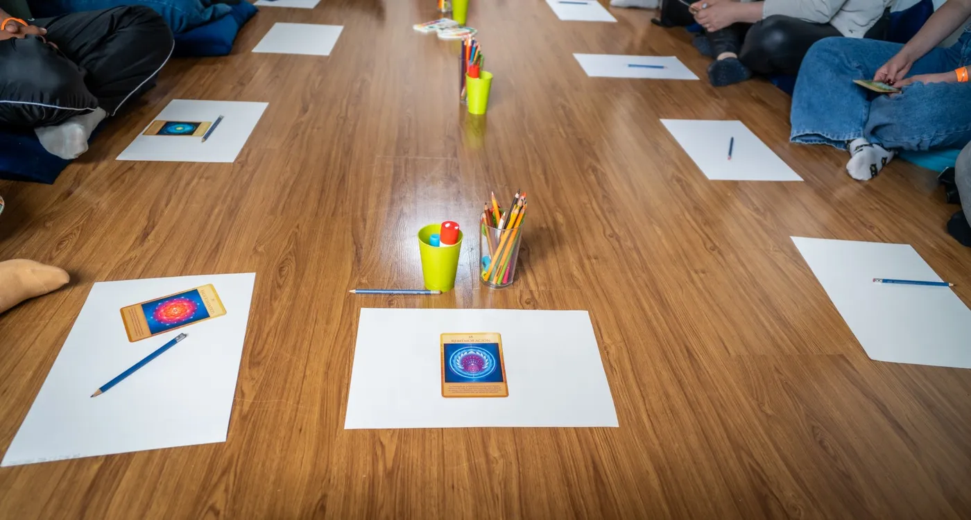 Hojas blancas con cartas de colores y vasos de lápices ordenados en círculo sobre el piso de un salón