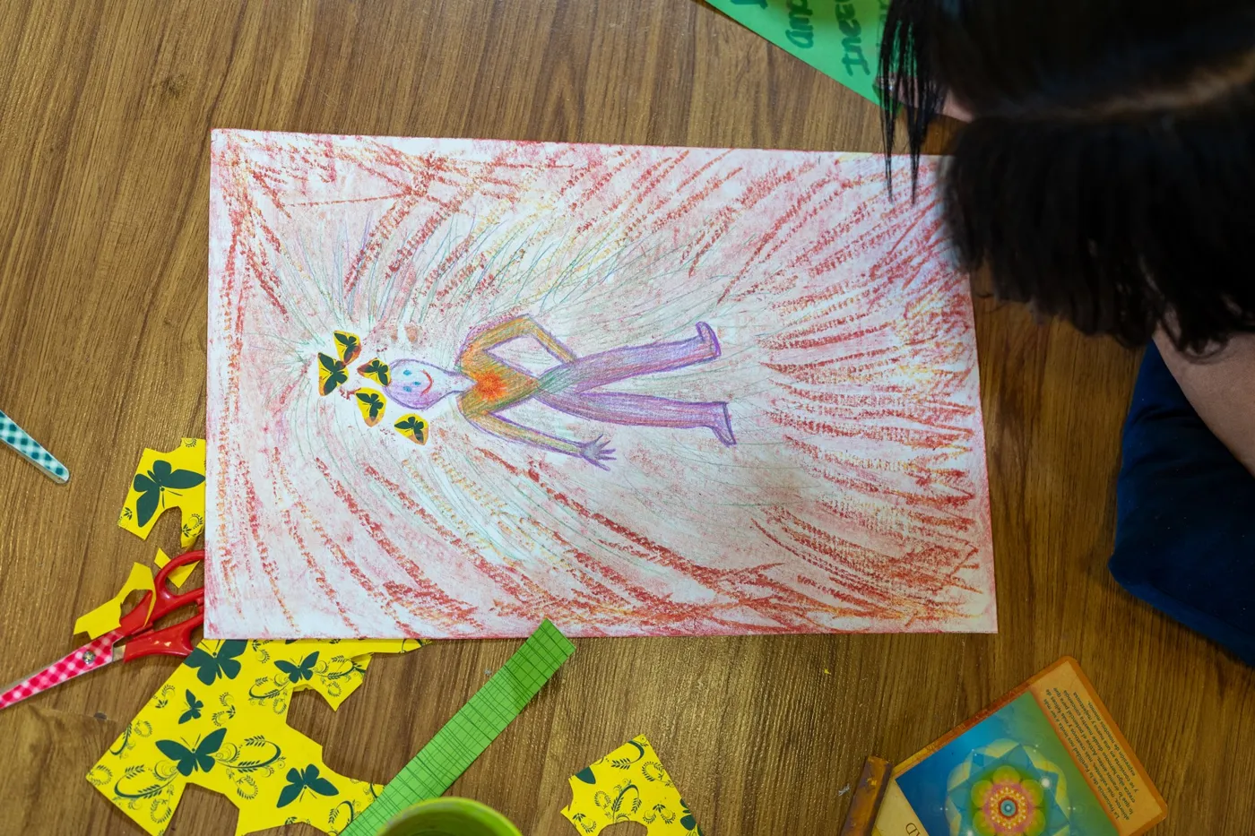 Un dibujo de una figura humana rodeada de trazos rojos, con mariposas de papel alrededor